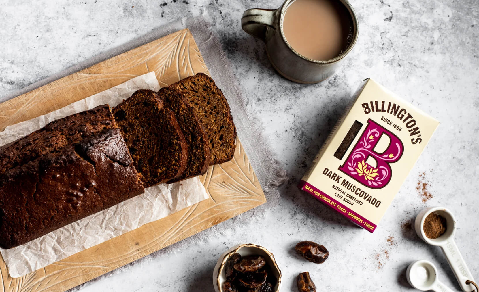 A sliced loaf cake and a coffee on a tabletop, with baking ingredients nearby, including Billington's Dark Muscovado Sugar.