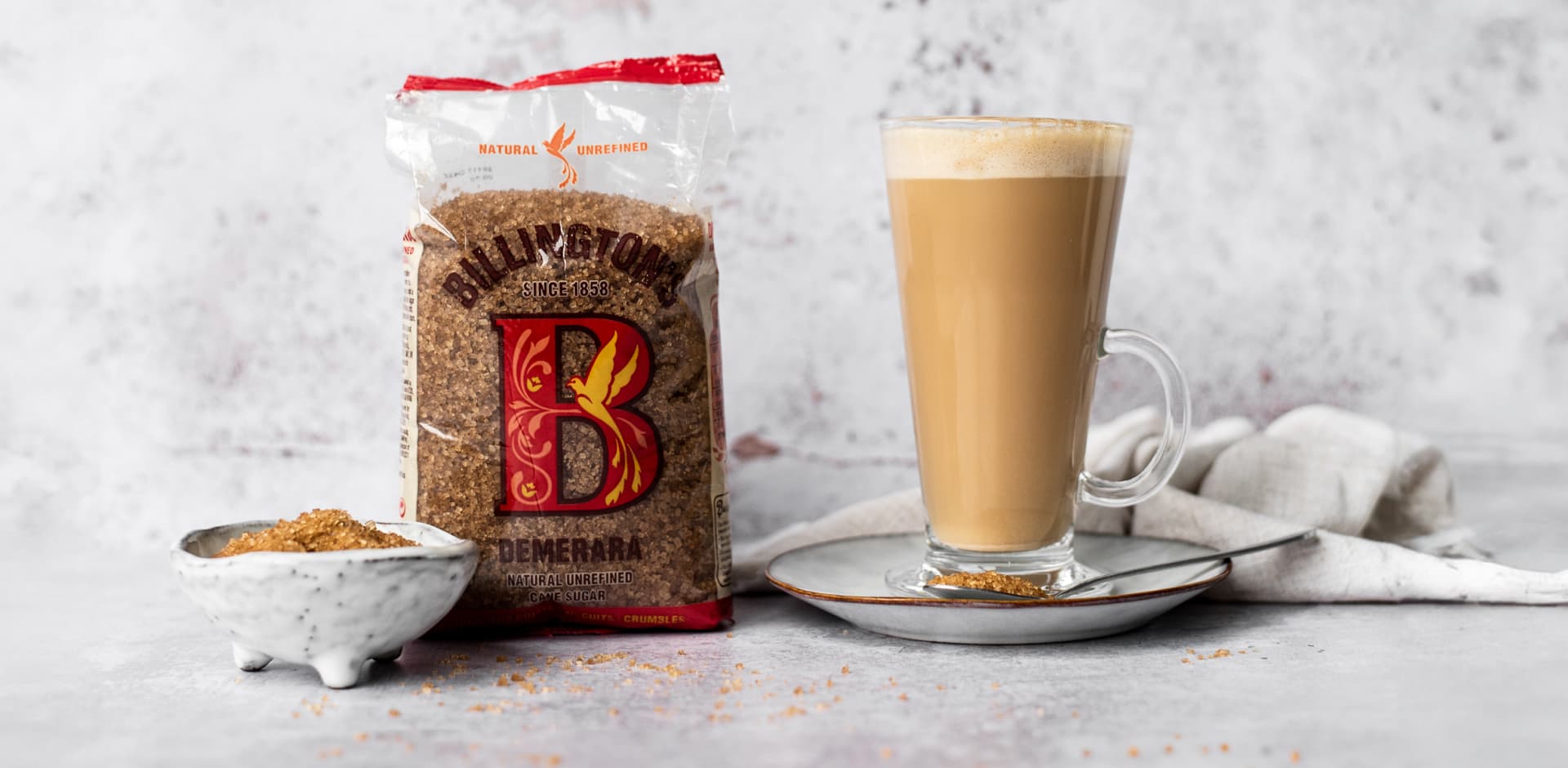 A pack of Billington's Demerara sugar stands next to a frothy latte in a tall glass.